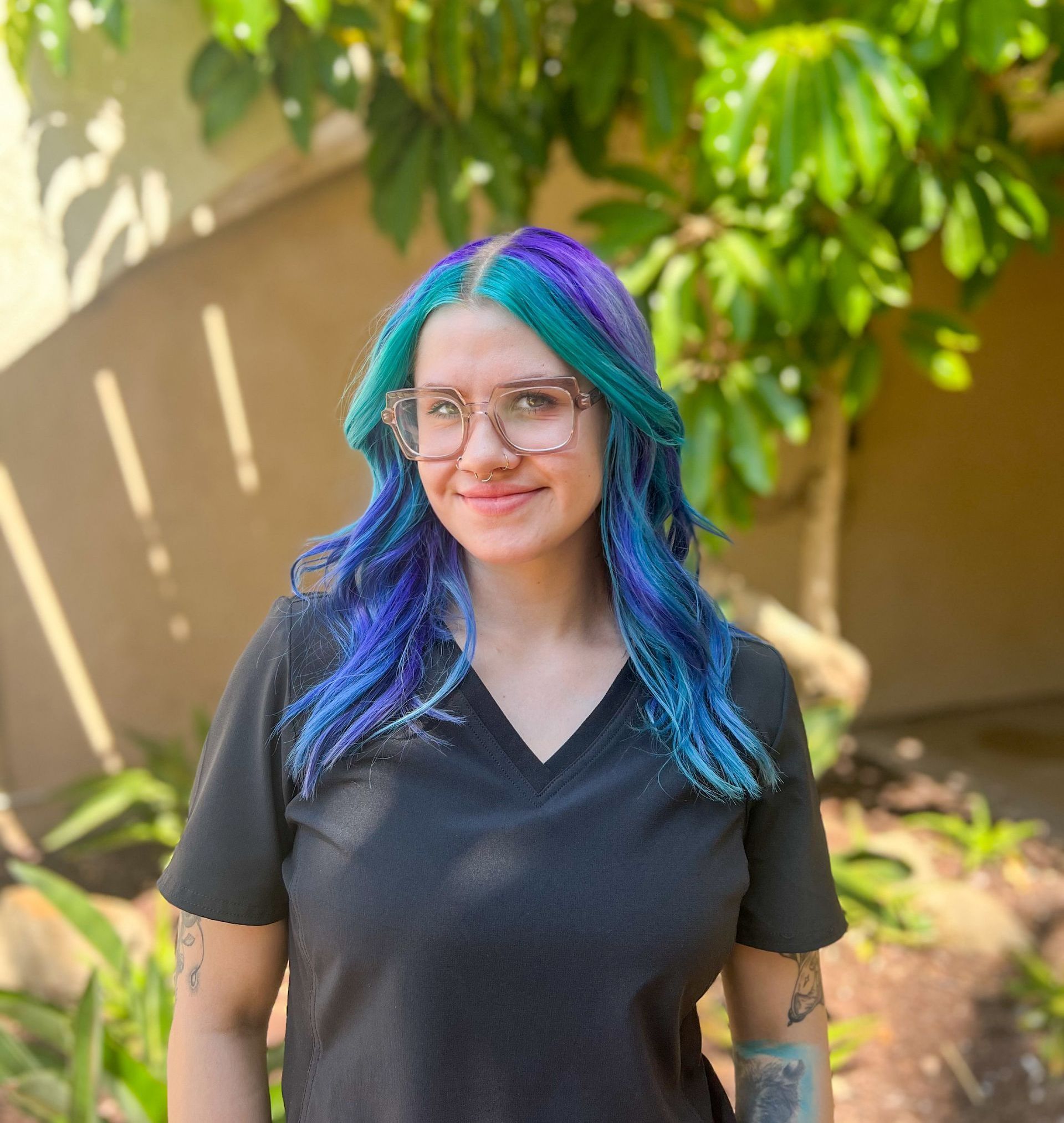 Woman with blue/purple hair, glasses, black shirt smiling in front of green foliage.