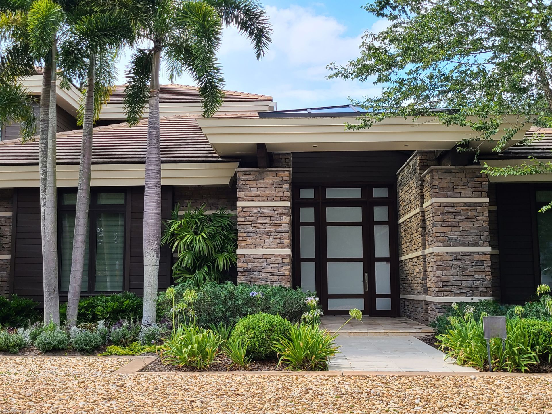 Modern home exterior with stone accents, glass door, and palm trees in sunny setting.