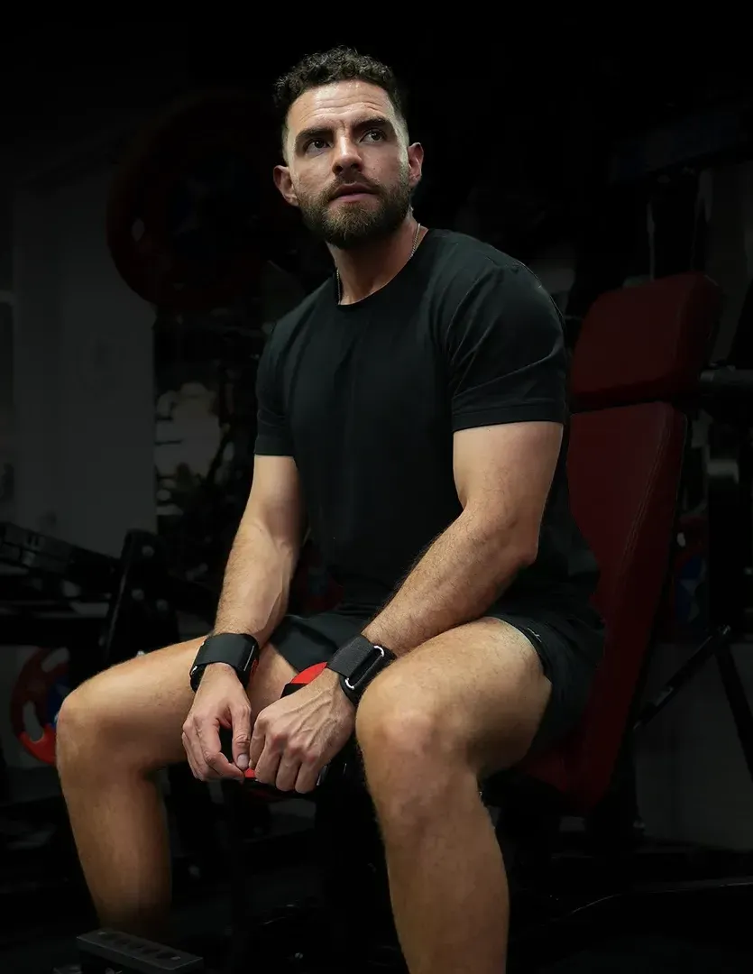 Man in black workout attire, seated on gym equipment, looking up with a determined expression. LEVAFIT