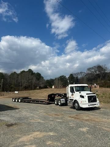 A white semi-truck with an attached lowboy trailer parked on a gravel lot under a cloudy blue sky.