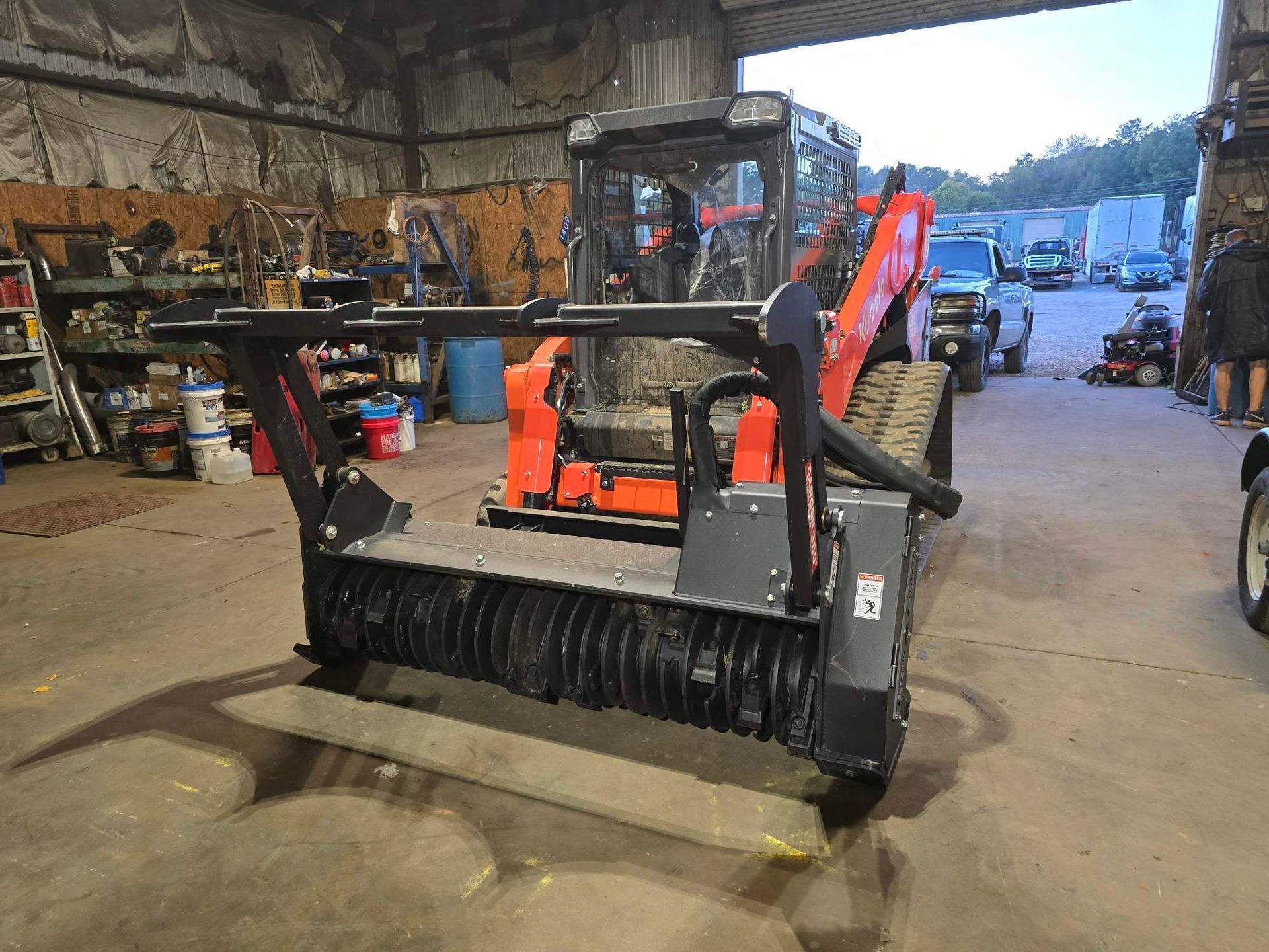 A black forestry mulcher attachment sits in the foreground, connected to an orange skid-steer loader inside a workshop.