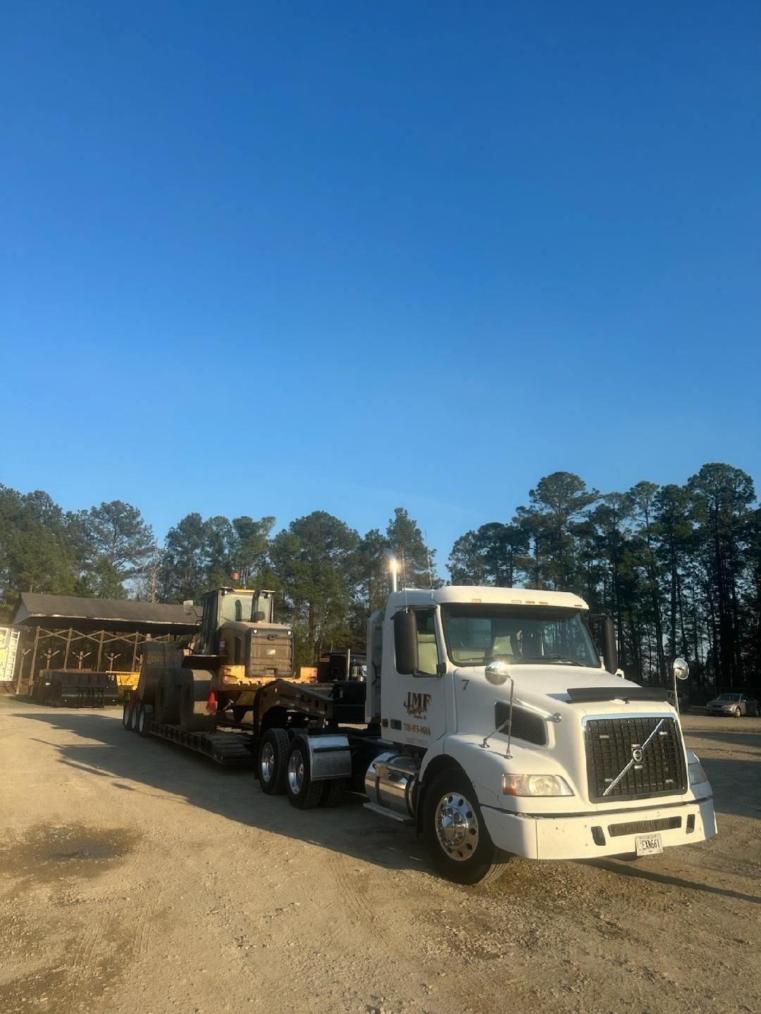 A white Volvo semi-truck hauls yellow heavy construction equipment on a flatbed trailer in a gravel lot under clear skies.