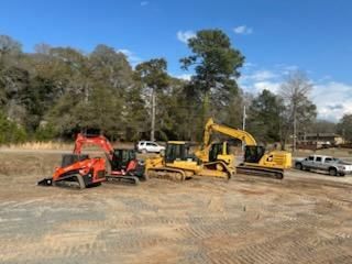 A red skid steer, yellow bulldozer, and yellow excavator parked on a dirt construction site with vehicles in the back.
