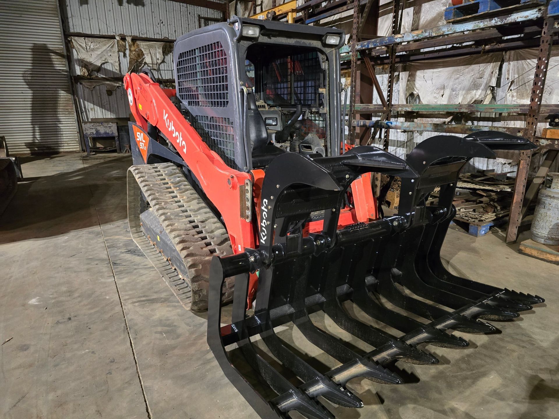 An orange and grey tracked skid steer loader with a large black grapple attachment, parked inside a warehouse.
