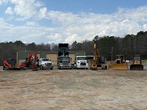 A line of heavy construction equipment and commercial trucks parked on a dirt lot in front of a metal maintenance building.