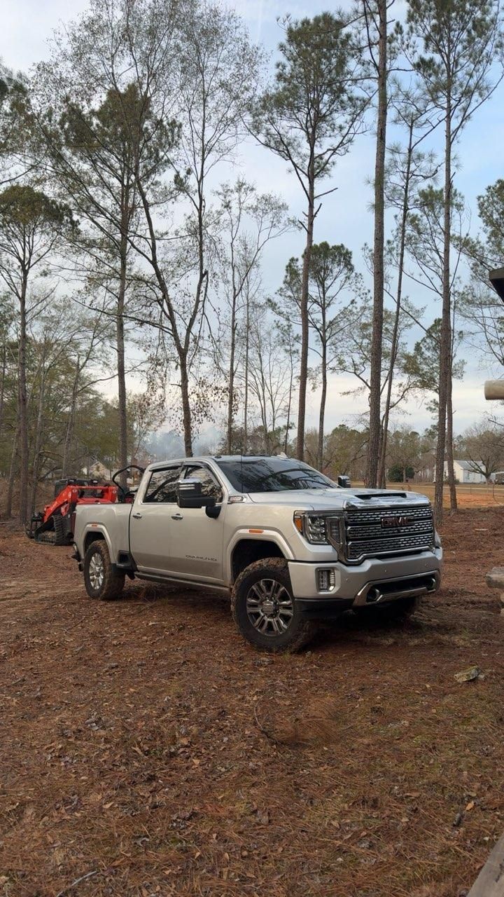 A light-colored heavy-duty pickup truck parked in a wooded area with an orange tractor visible in the background.