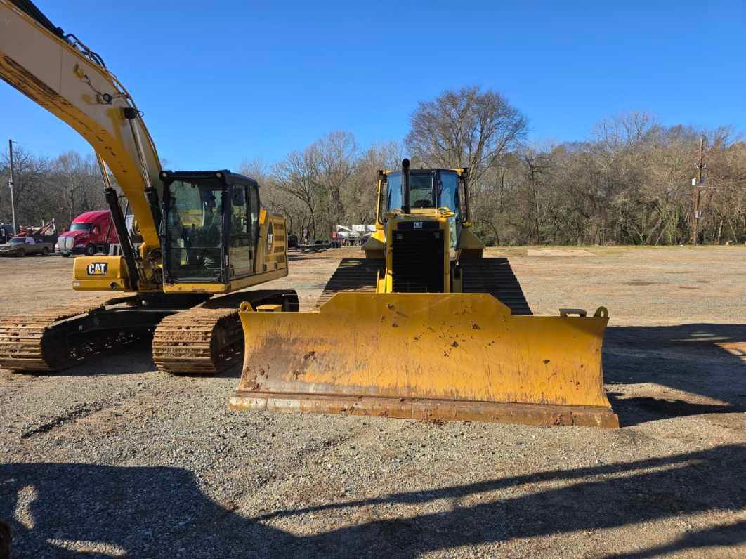 A yellow CAT excavator and a bulldozer sit parked side-by-side on a gravel lot under a clear blue sky.