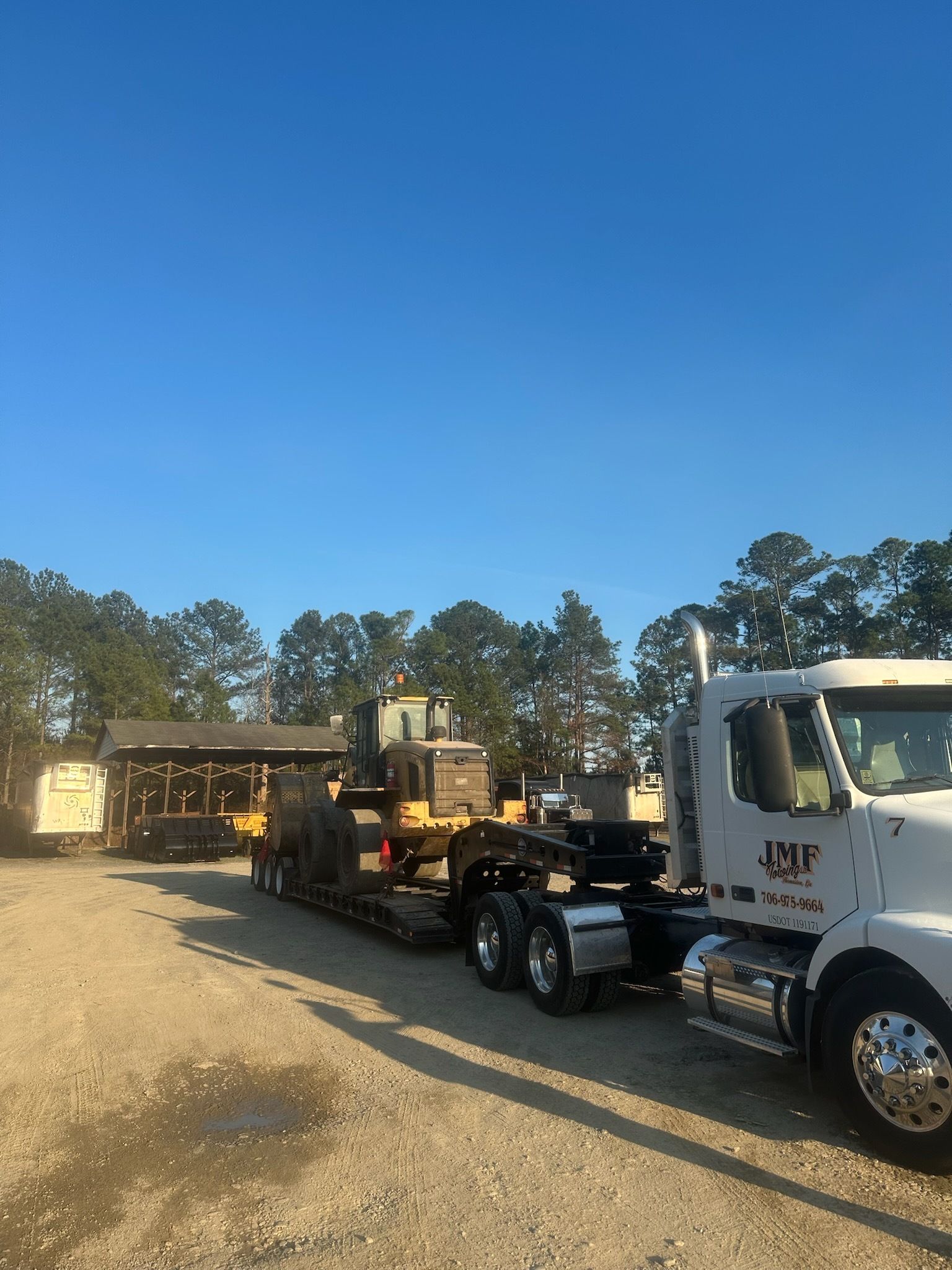 A white JMF truck towing a yellow construction vehicle on a flatbed trailer in a sunny, tree-lined gravel lot.