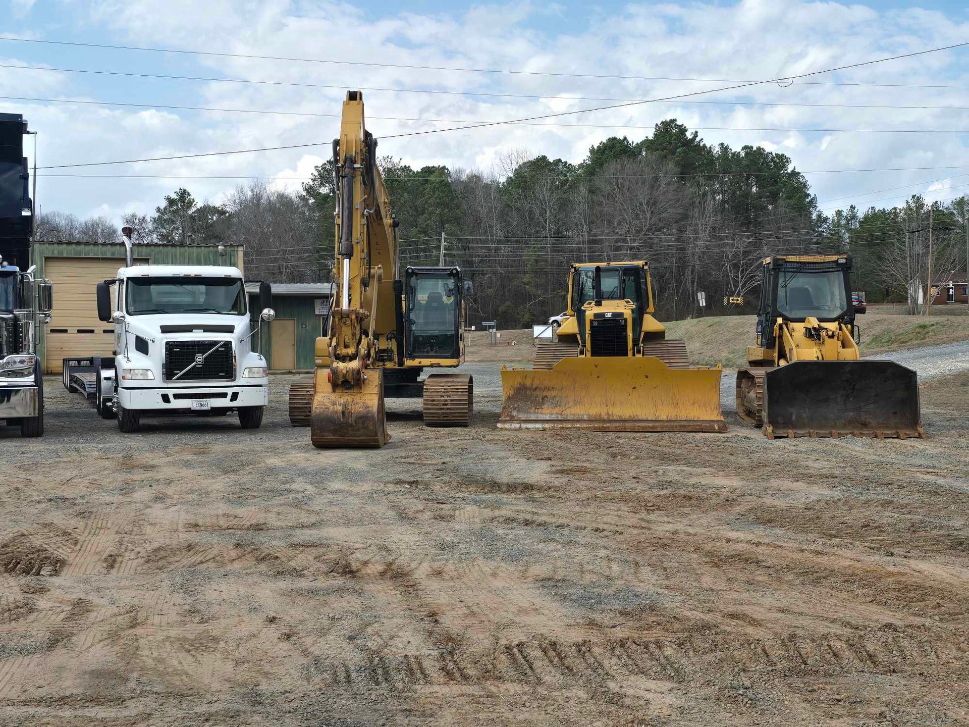 A white Volvo truck, a tall yellow excavator, and two yellow bulldozers parked on a dirt lot with trees in the background.