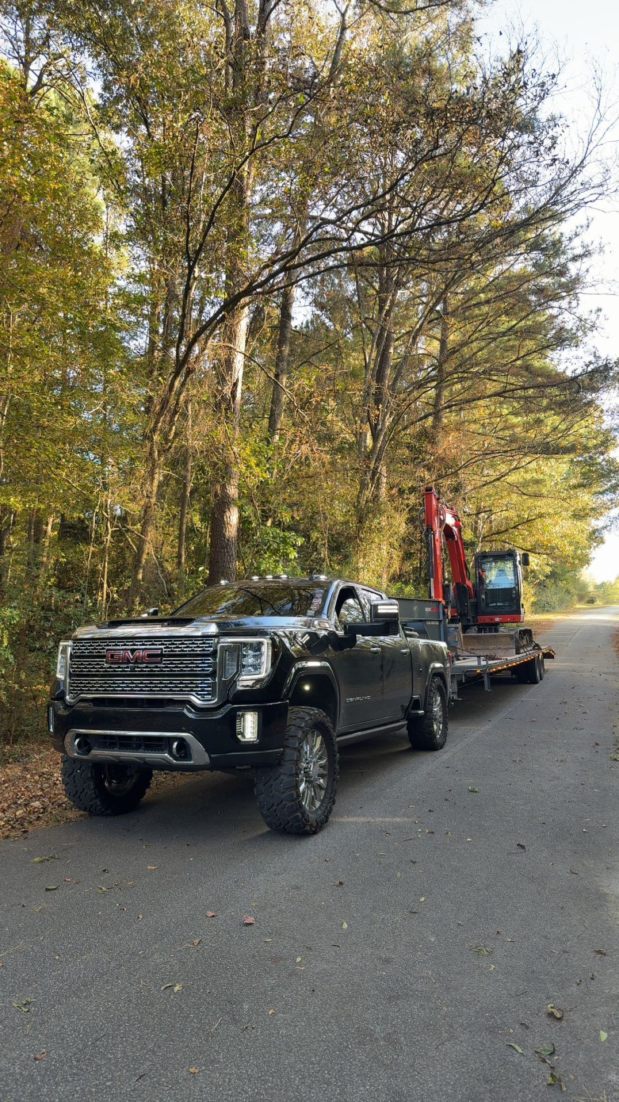 A black GMC pickup truck towing a trailer with a red excavator on a wooded road.