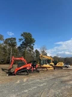 A red tracked skid steer and two yellow excavators parked in a line on a dirt construction lot under a blue sky.