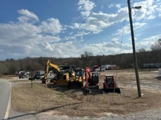 Construction equipment including a bulldozer and excavators parked on a dirt lot under a blue sky with clouds.