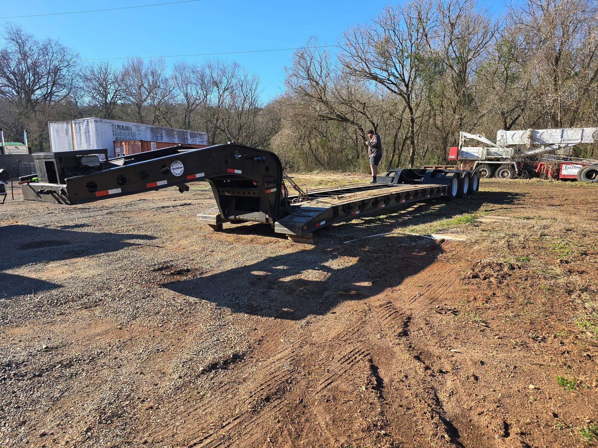 A black lowboy semi-trailer sits on a dirt lot with a person standing near it under a clear blue sky.