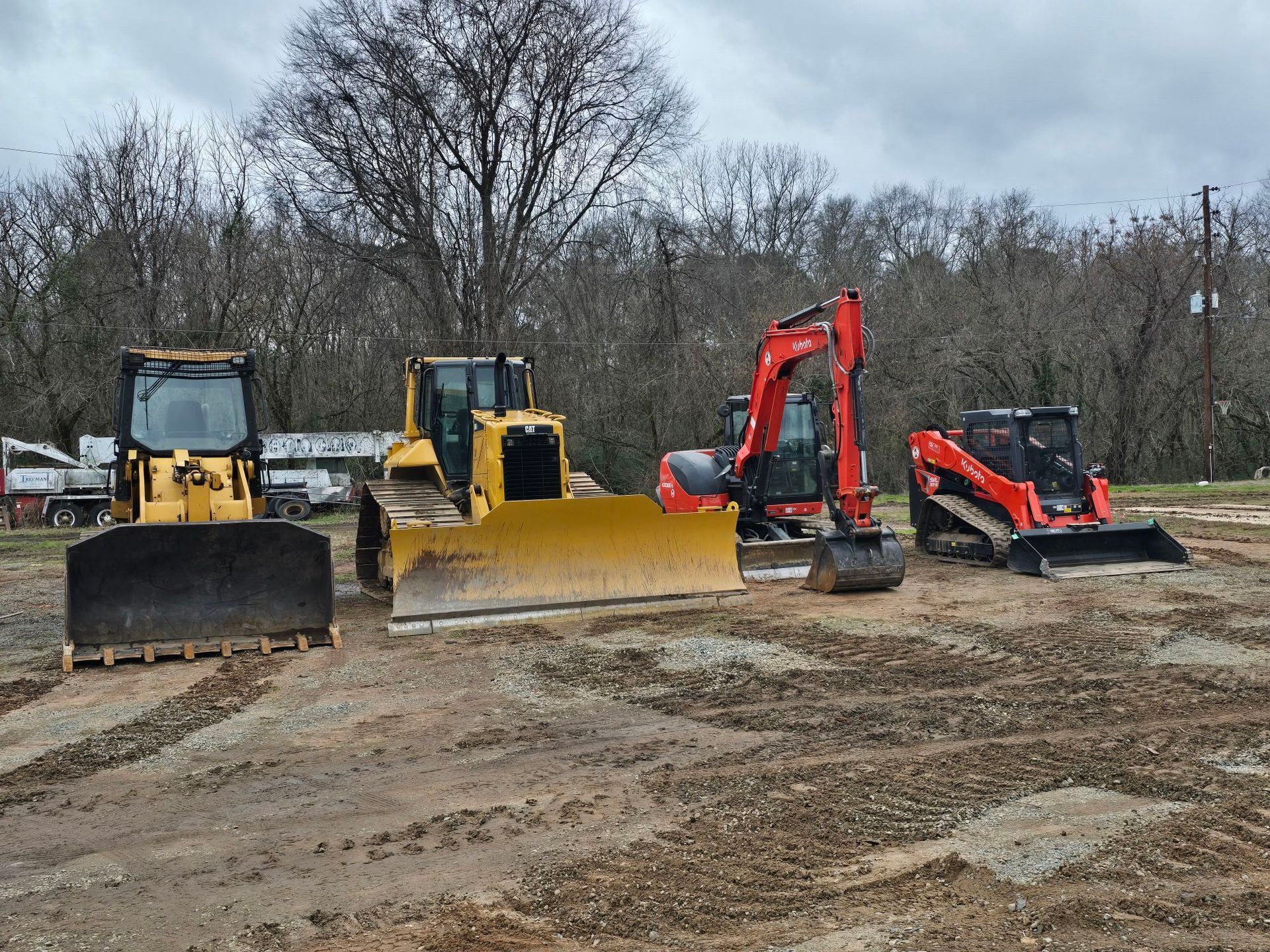 Four pieces of heavy construction equipment, including bulldozers, an excavator, and a skid steer, parked in a field.