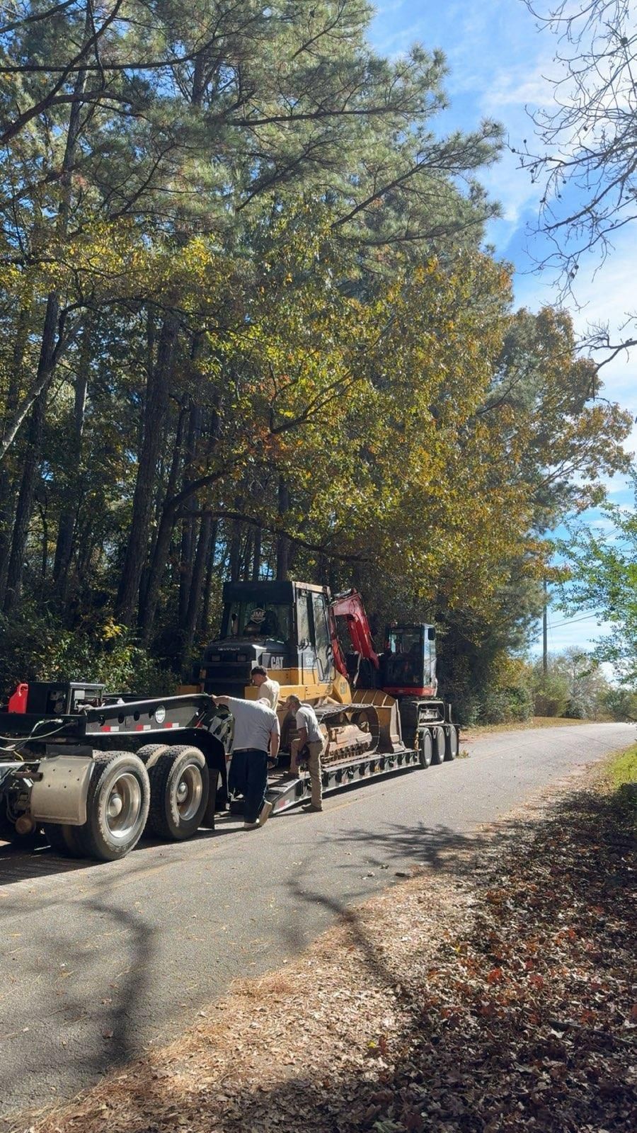 A flatbed truck parked on a rural road, loaded with heavy yellow construction equipment, with two workers standing nearby.