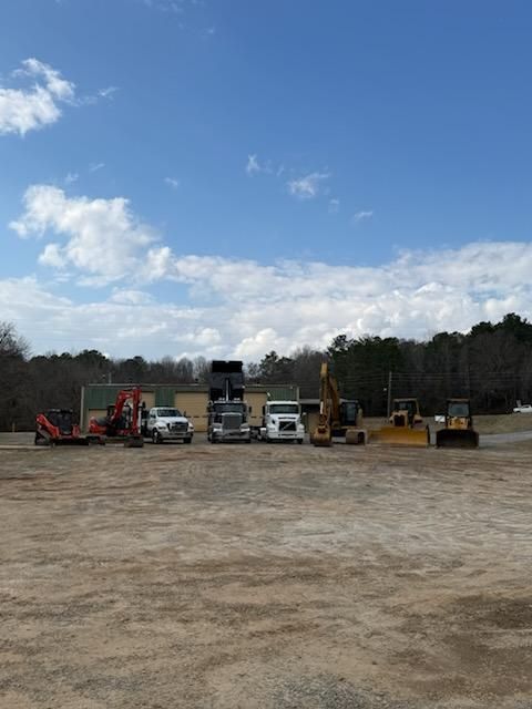 A line of construction vehicles and trucks parked in a dirt lot under a blue sky with clouds.