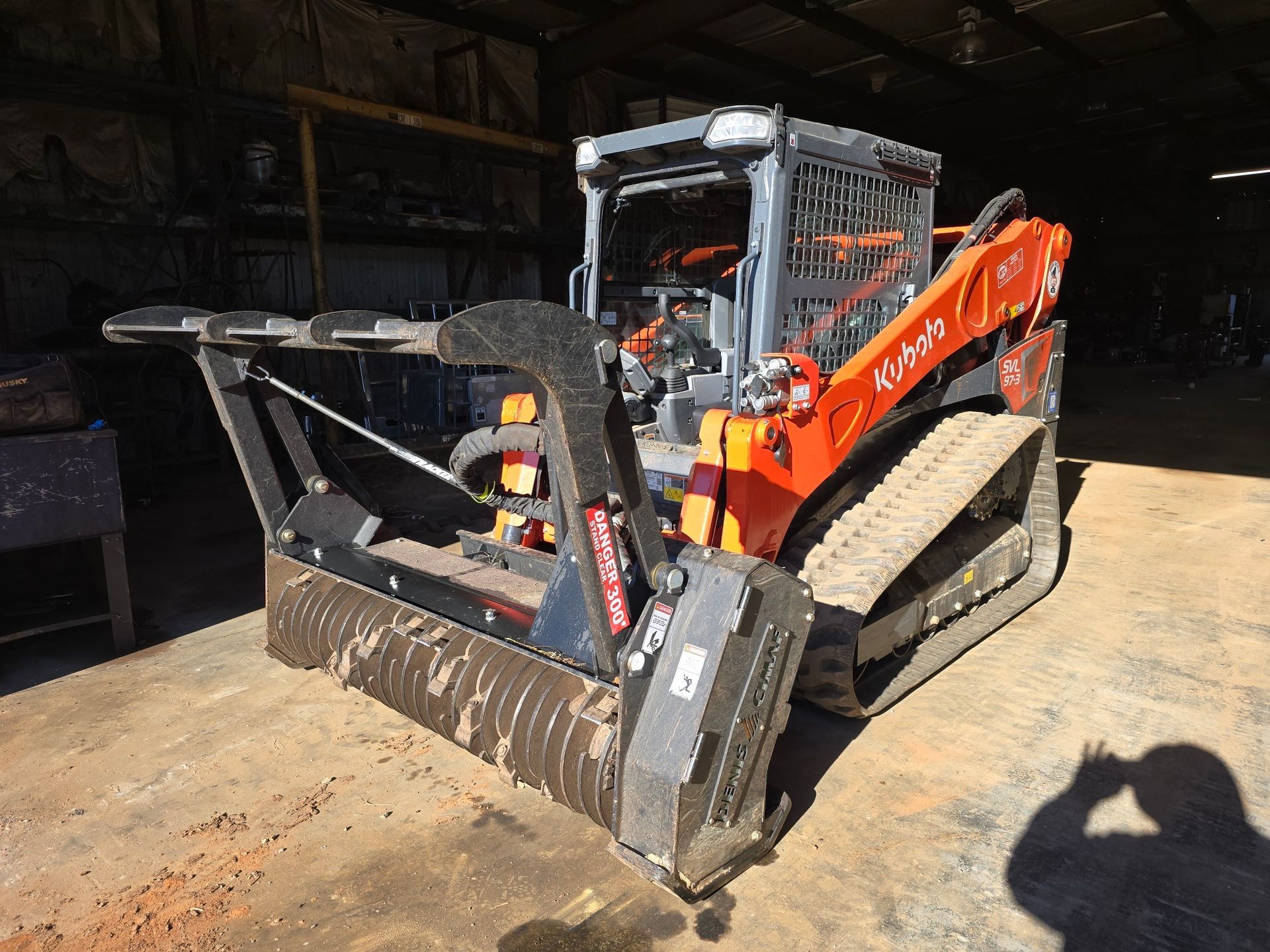 A Kubota compact track loader with a large forestry mulcher attachment parked inside a workshop.