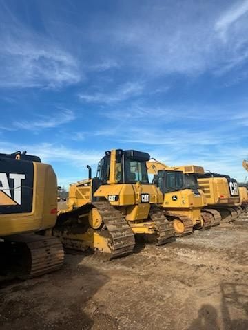 A row of yellow construction bulldozers parked in a dirt lot under a bright blue sky.