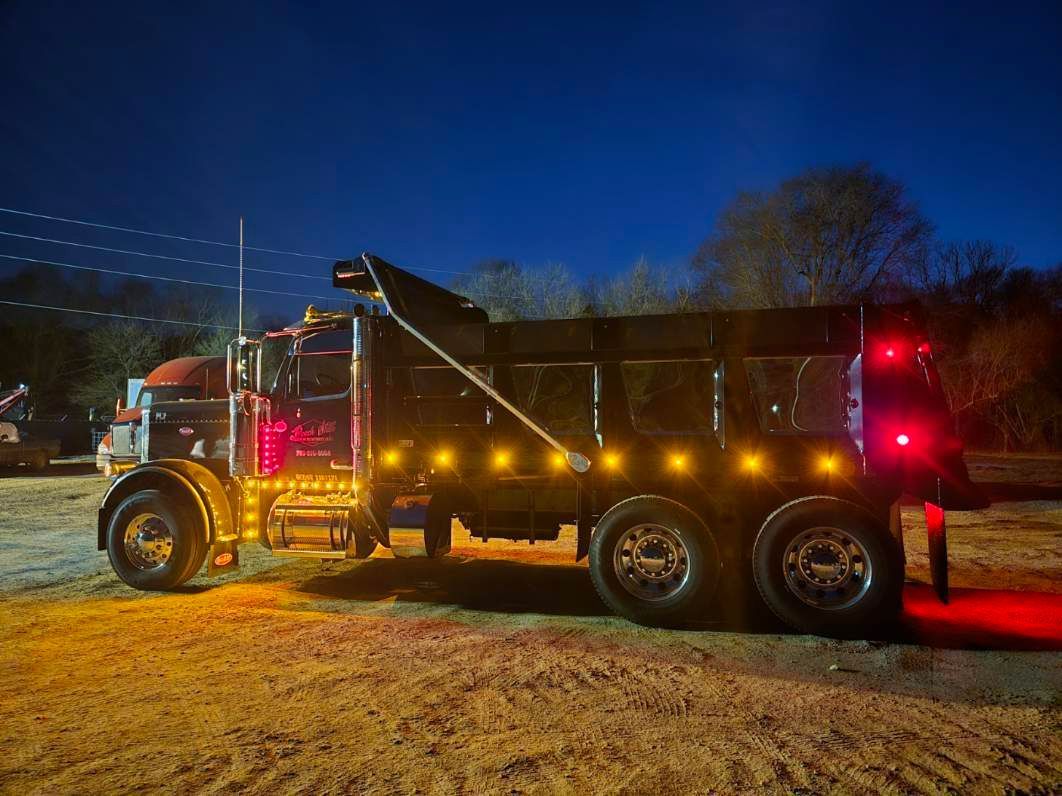 A black dump truck parked on a dirt lot at night, illuminated by yellow side lights and bright red rear brake lights.