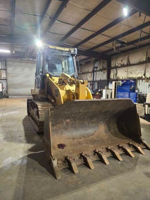 A yellow Caterpillar track loader with a large front bucket parked inside a warehouse with metal ceiling beams.