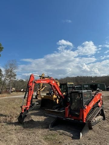 Two orange Kubota construction machines, a mini excavator and a compact track loader, parked on a dirt lot under a blue sky.