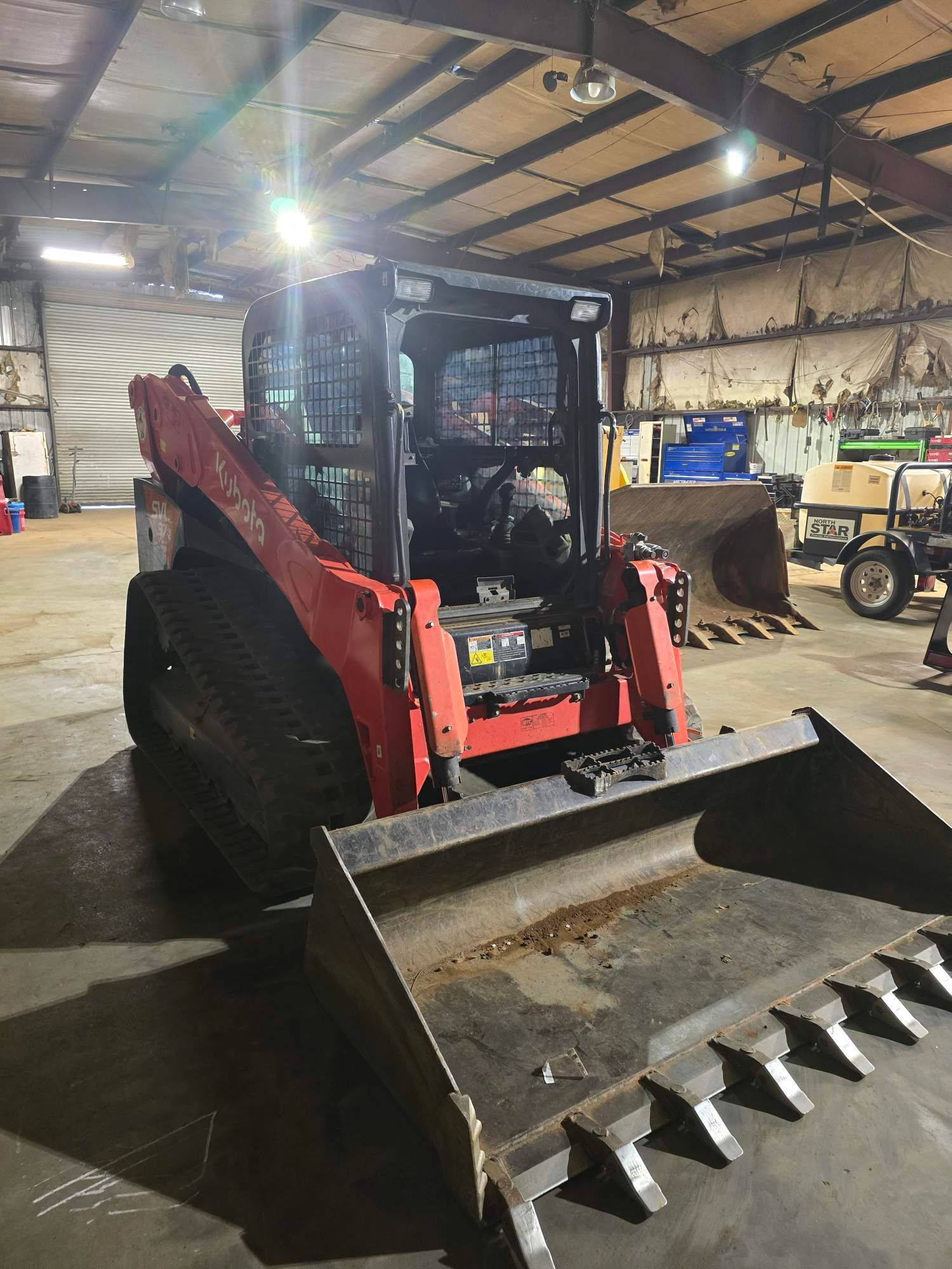 A red compact track loader with a toothed bucket sits inside a large, industrial warehouse.