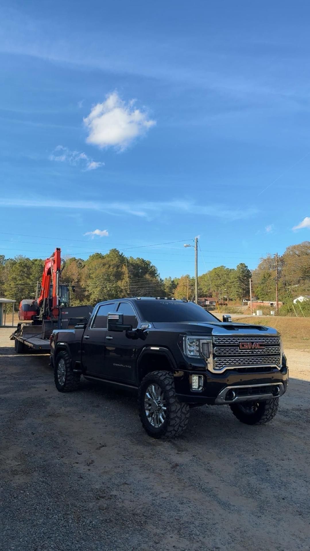 A black GMC pickup truck with lifted tires is parked on a gravel lot, towing an orange excavator on a trailer.