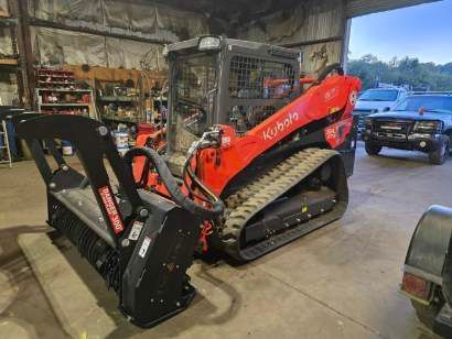 Orange Kubota compact track loader with a black forestry mulcher attachment parked inside a workshop.