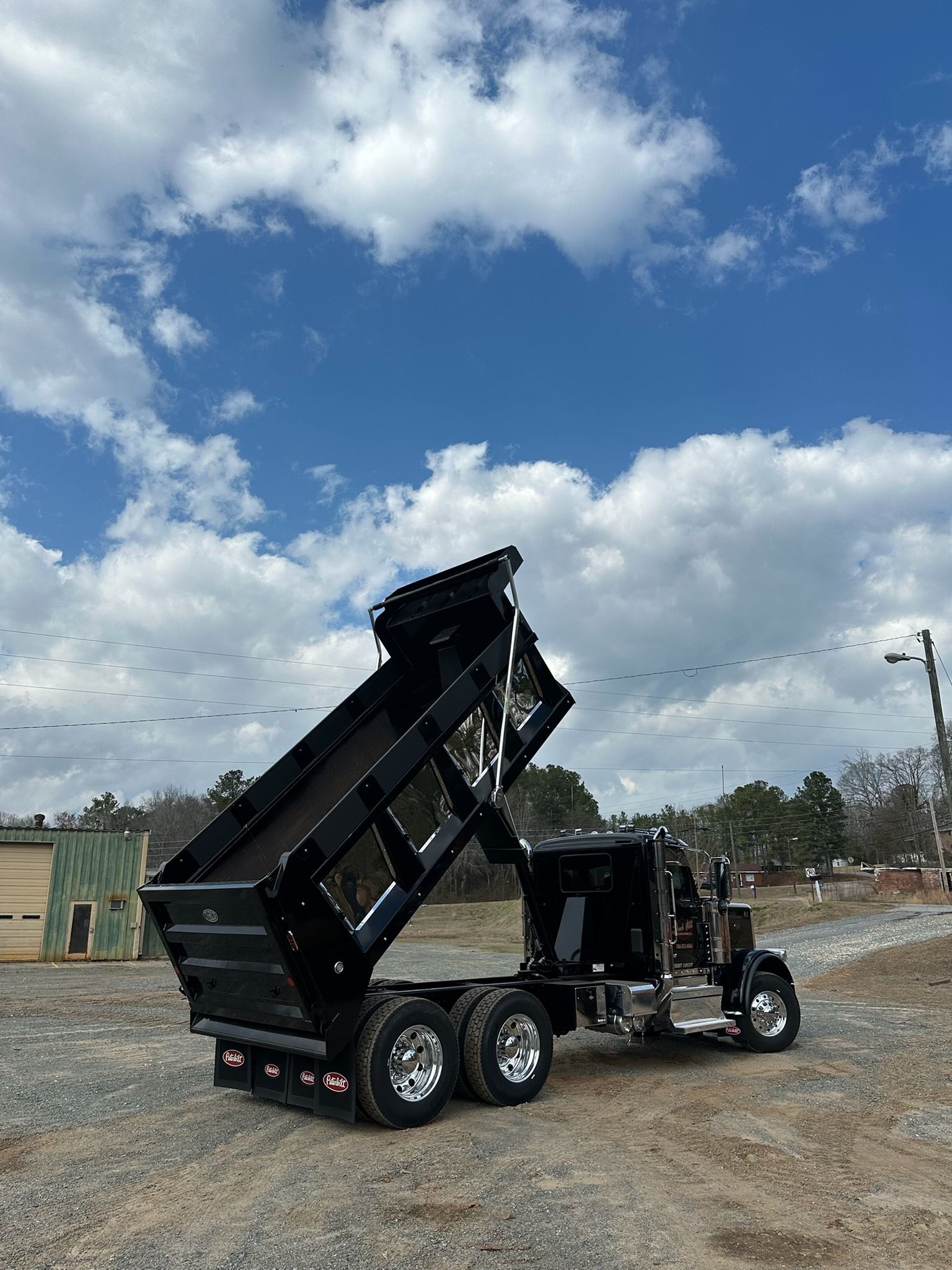 A black dump truck with its bed fully raised, parked on a gravel lot under a blue sky with scattered clouds.