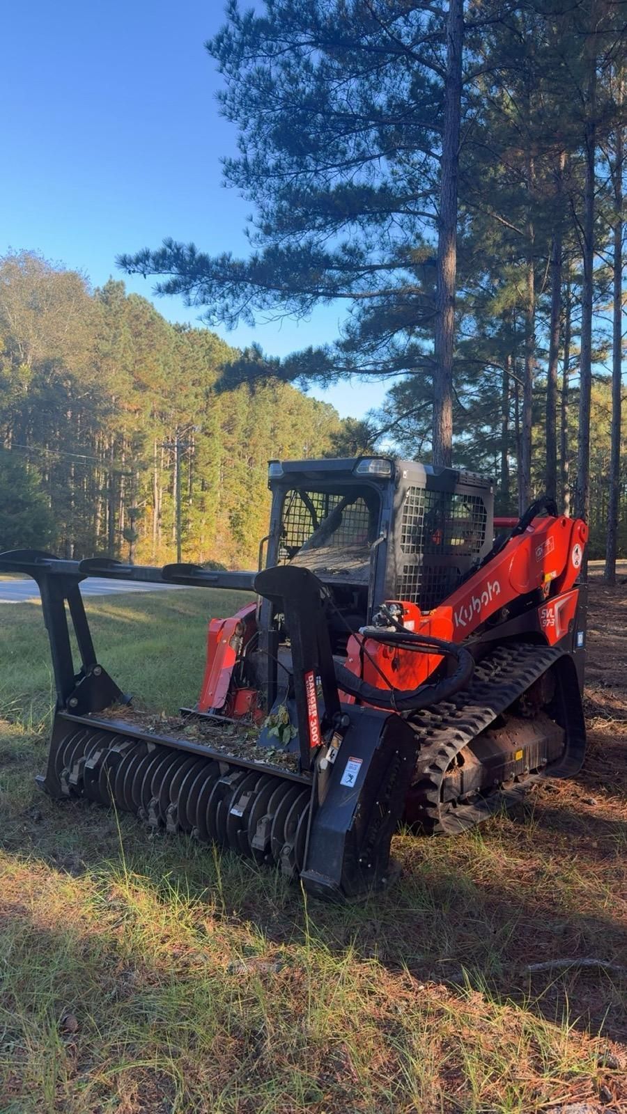 An orange Kubota track loader with a mulcher attachment sits in a grassy area near a line of trees on a sunny day.