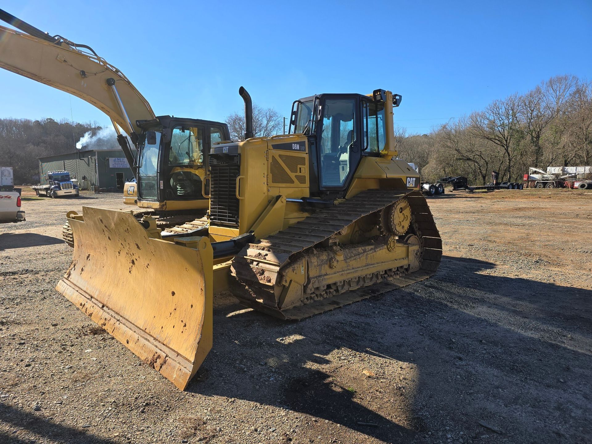 A white semi-truck with a lowboy trailer parked on a gravel lot under a bright, cloudy blue sky.