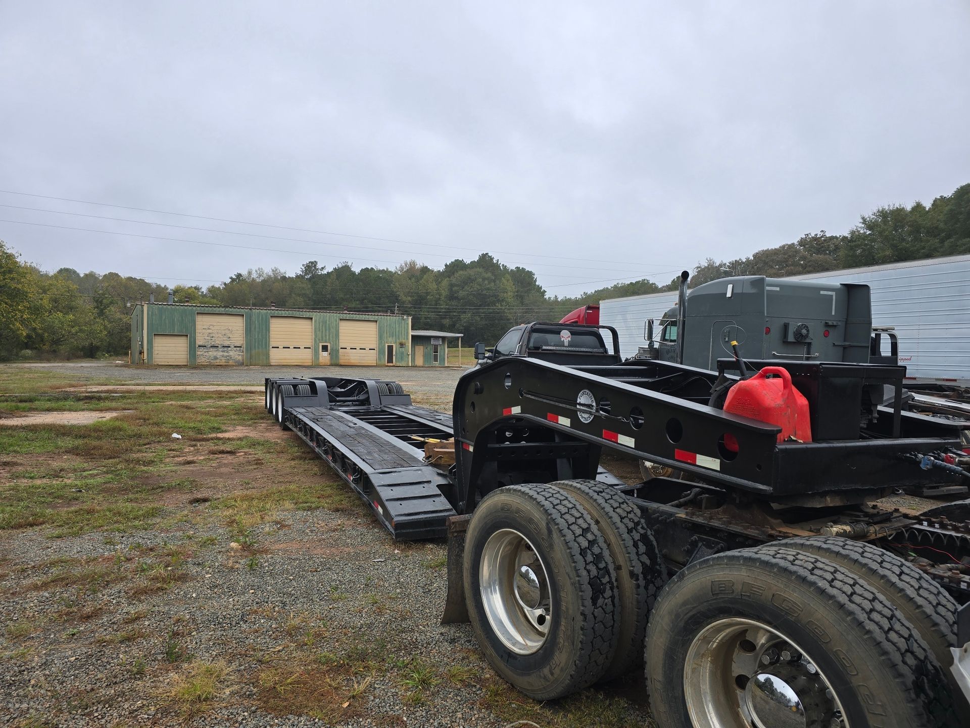A semi-truck with a lowboy trailer parked on a gravel lot in front of a metal warehouse.