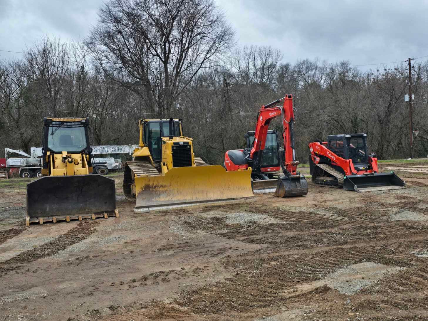 Four pieces of heavy construction equipment, including bulldozers and an excavator, parked on a dirt lot.