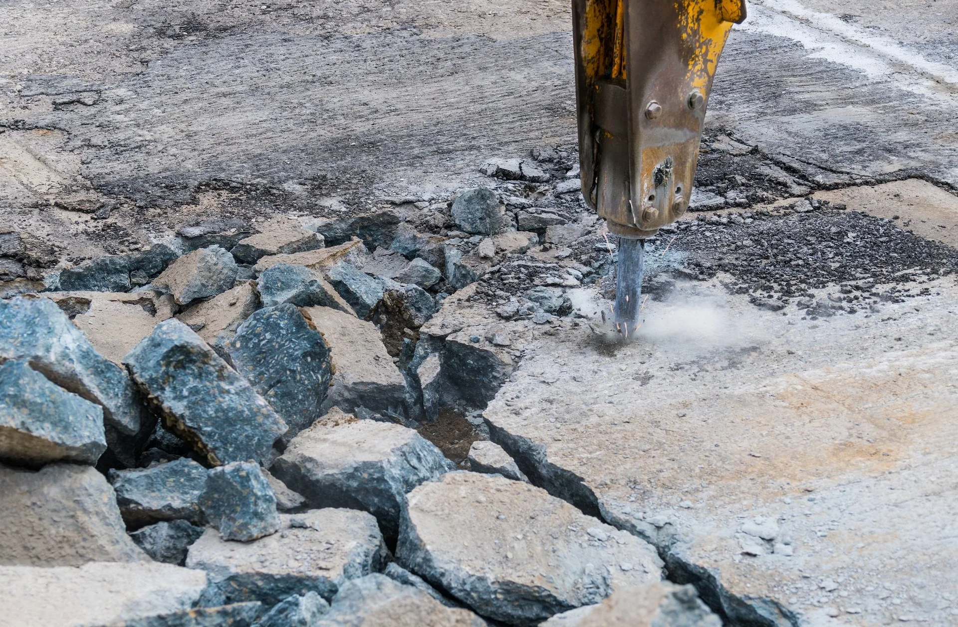 A heavy hydraulic hammer attachment on an excavator breaks up old pavement and rocks on a construction site.
