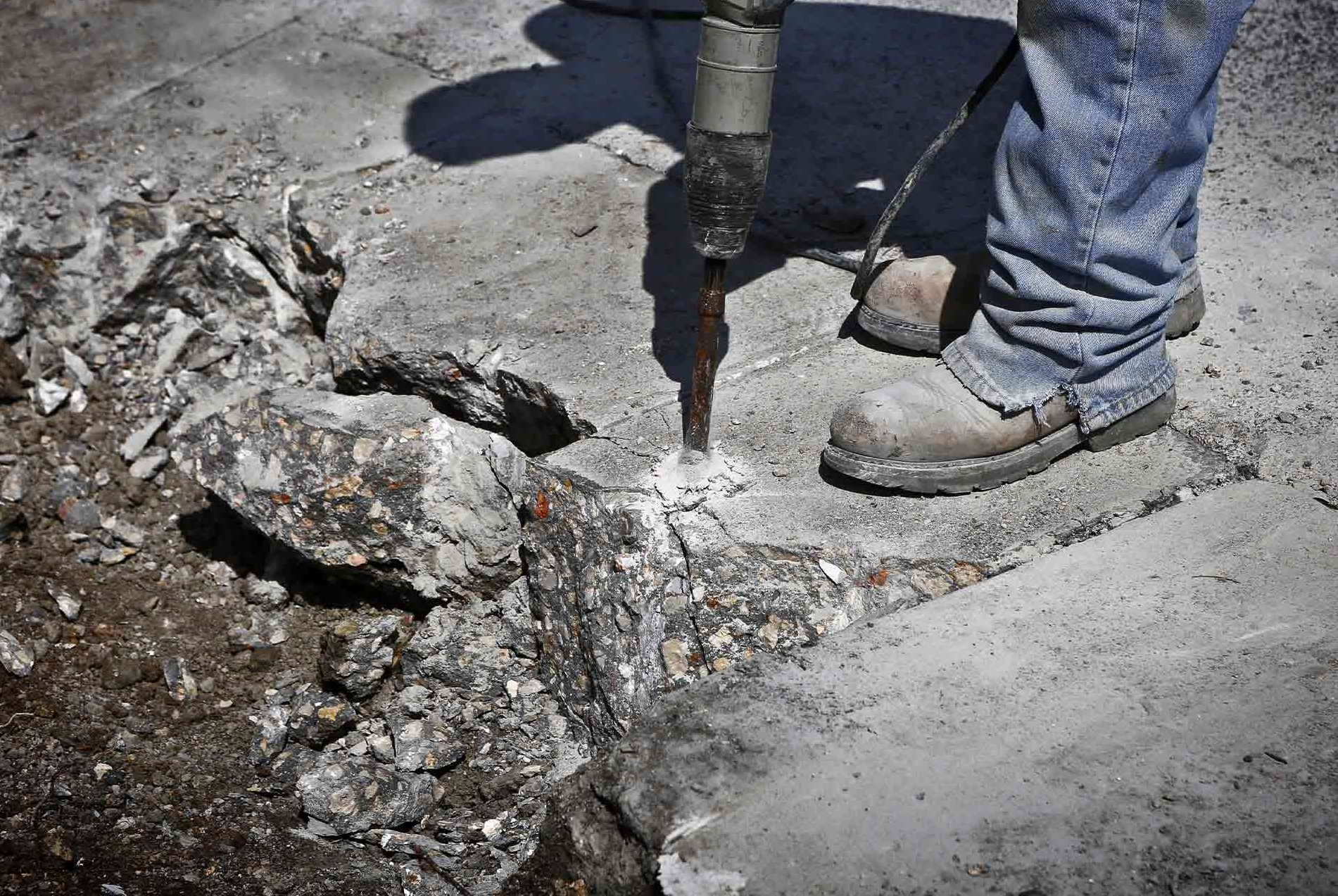 A worker in work boots uses a jackhammer to break up a concrete sidewalk.