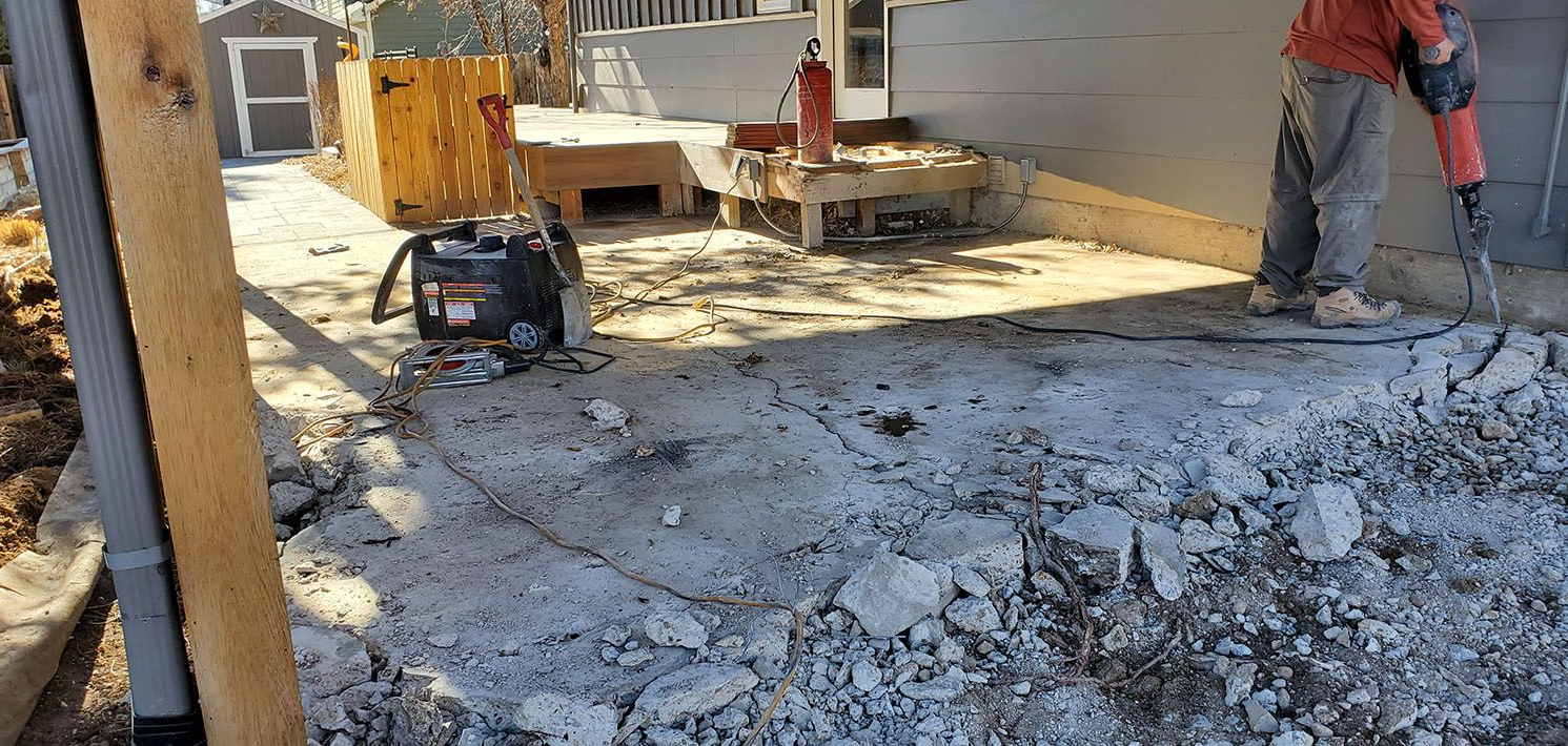 A worker uses a jackhammer to break up a concrete patio outside a house.