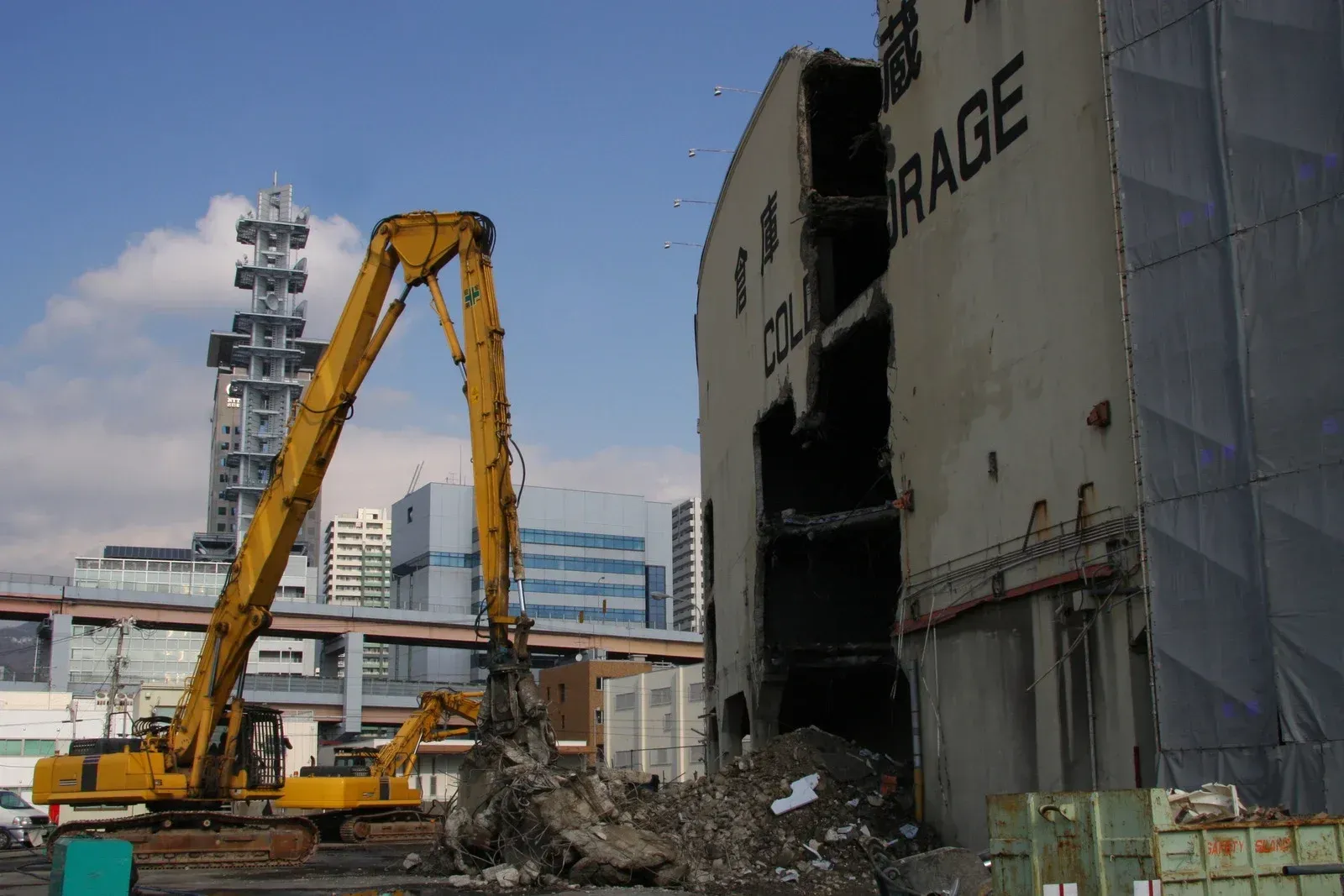 A yellow high-reach excavator tears down a multi-story concrete building at a construction site.