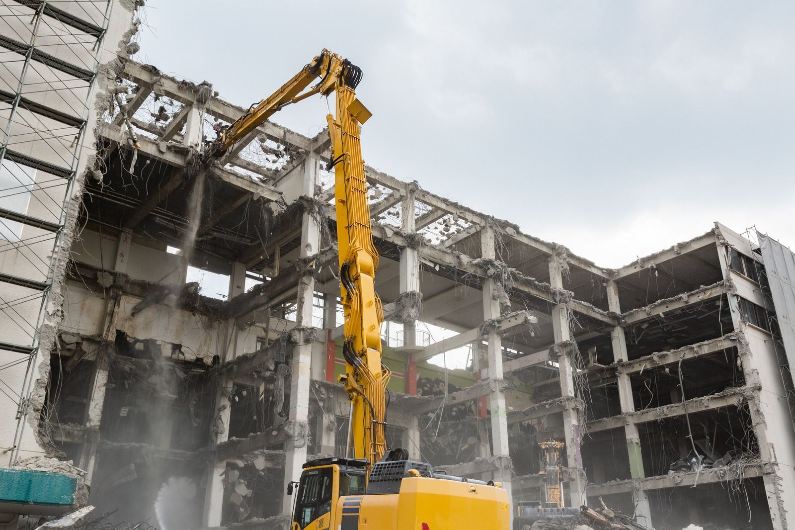A yellow high-reach demolition excavator works to tear down the concrete structure of a multi-story building.