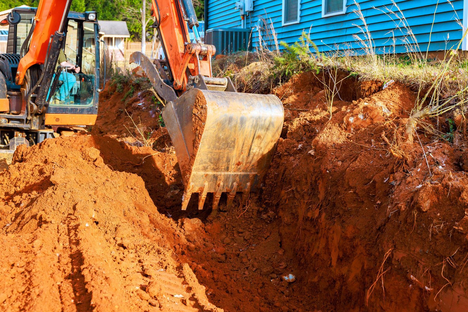 An orange excavator digging a deep trench in red clay soil next to a blue house.