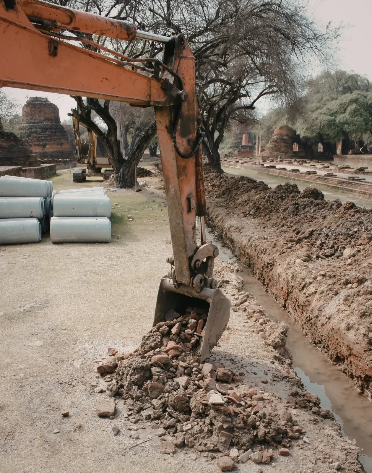 An orange excavator digging a narrow trench near historic brick ruins and trees.