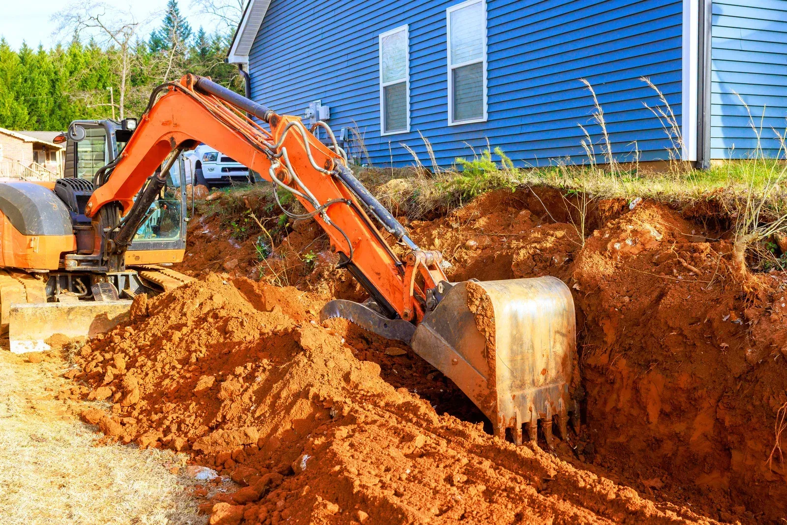 An orange excavator digging a trench in red dirt next to a blue house.