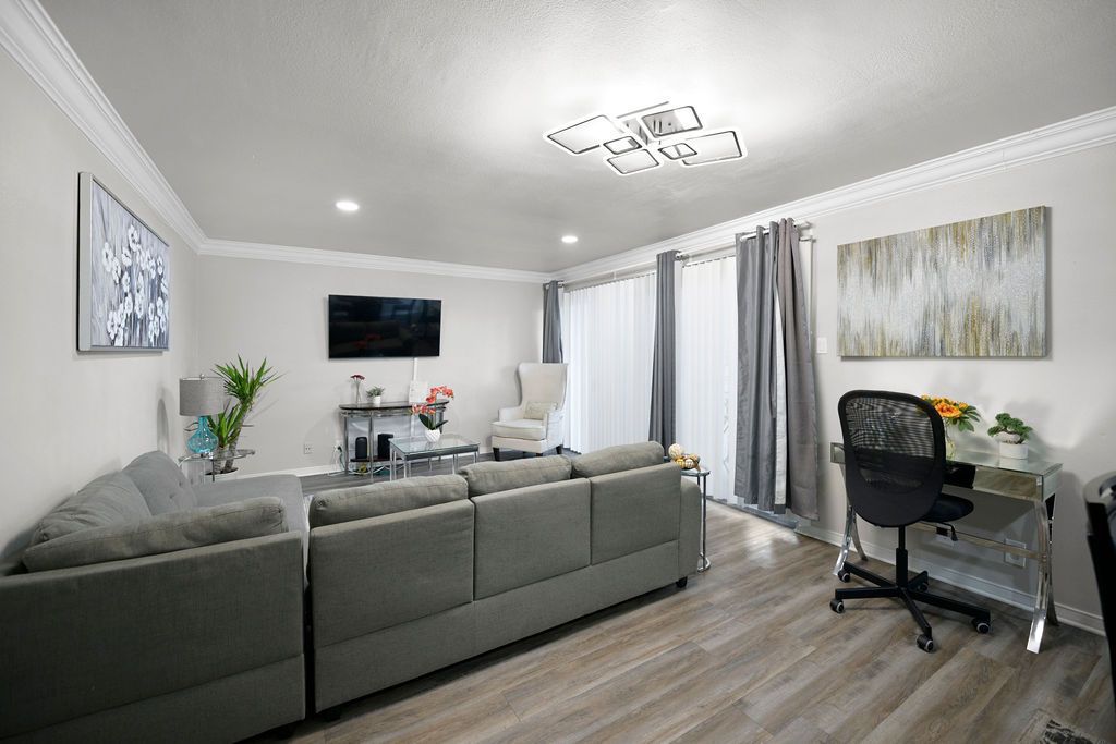 A modern living room with a gray sectional sofa, a TV, and a desk. Neutral colors dominate the space with light-colored walls, curtains, and flooring.