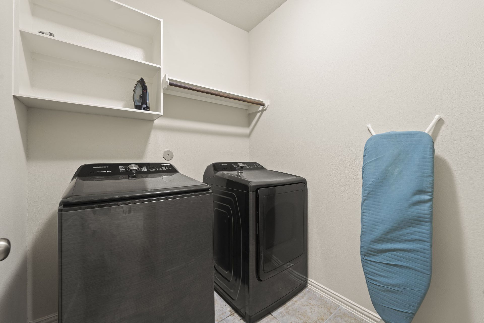 Laundry room with black washer and dryer, white shelves, and a blue ironing board.