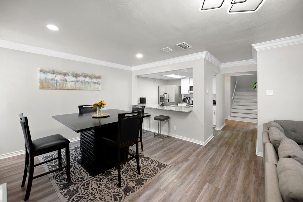 A dining room with a black square table and chairs, a bar area, and a staircase leading upwards. Grey walls and wood-look flooring.