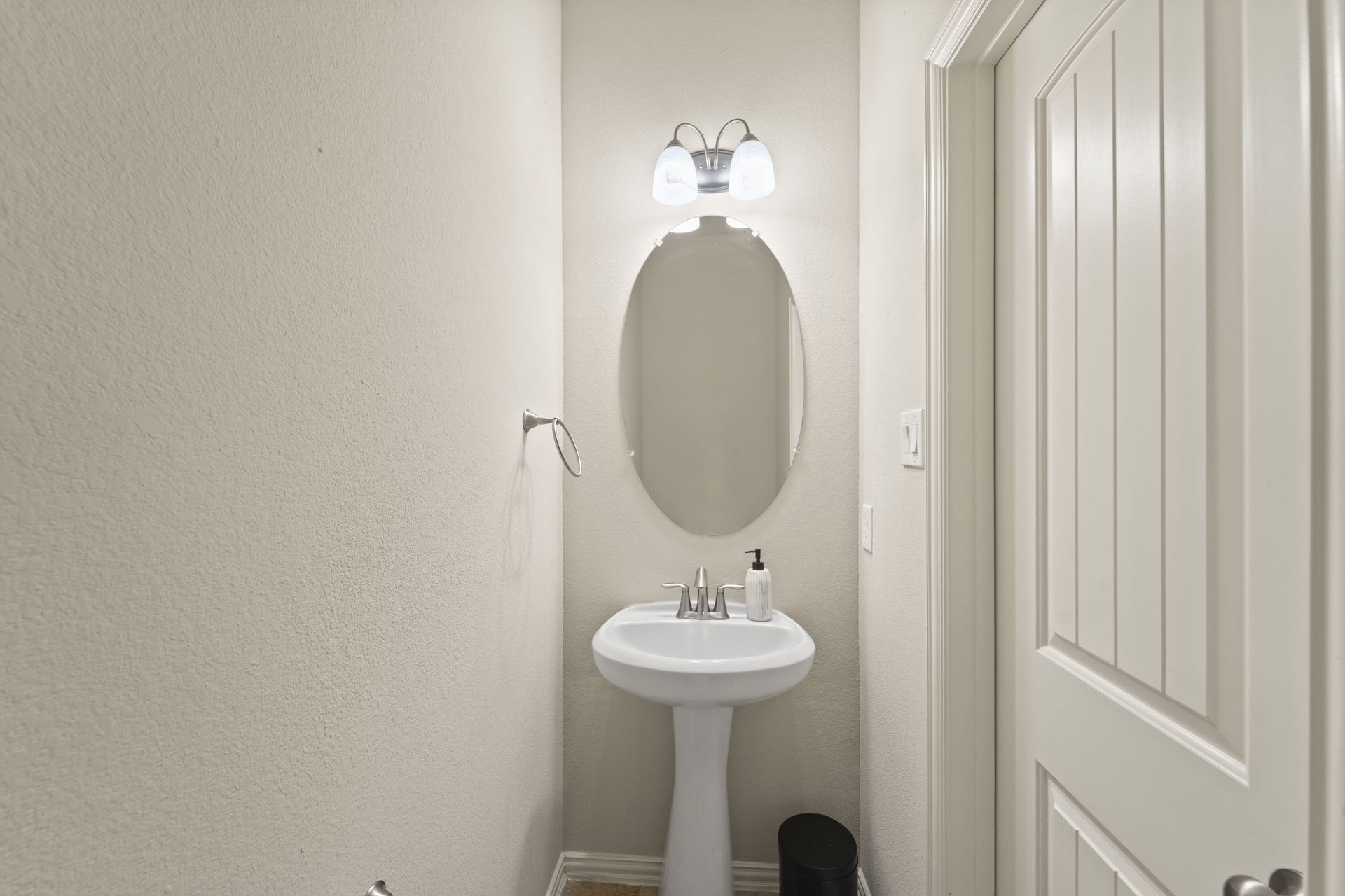 Small powder room with white pedestal sink, oval mirror, and white door.