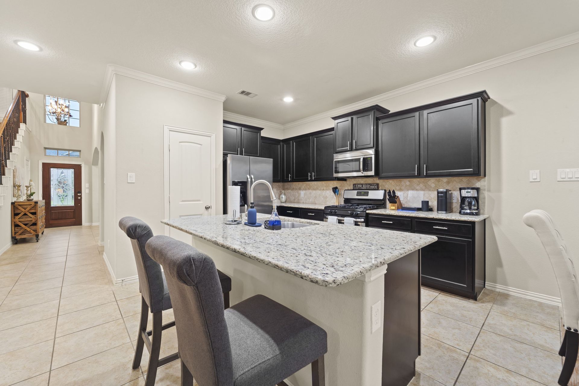 Kitchen with dark cabinets, granite countertops, and a breakfast bar with two gray upholstered stools. A hallway leads to the front door.