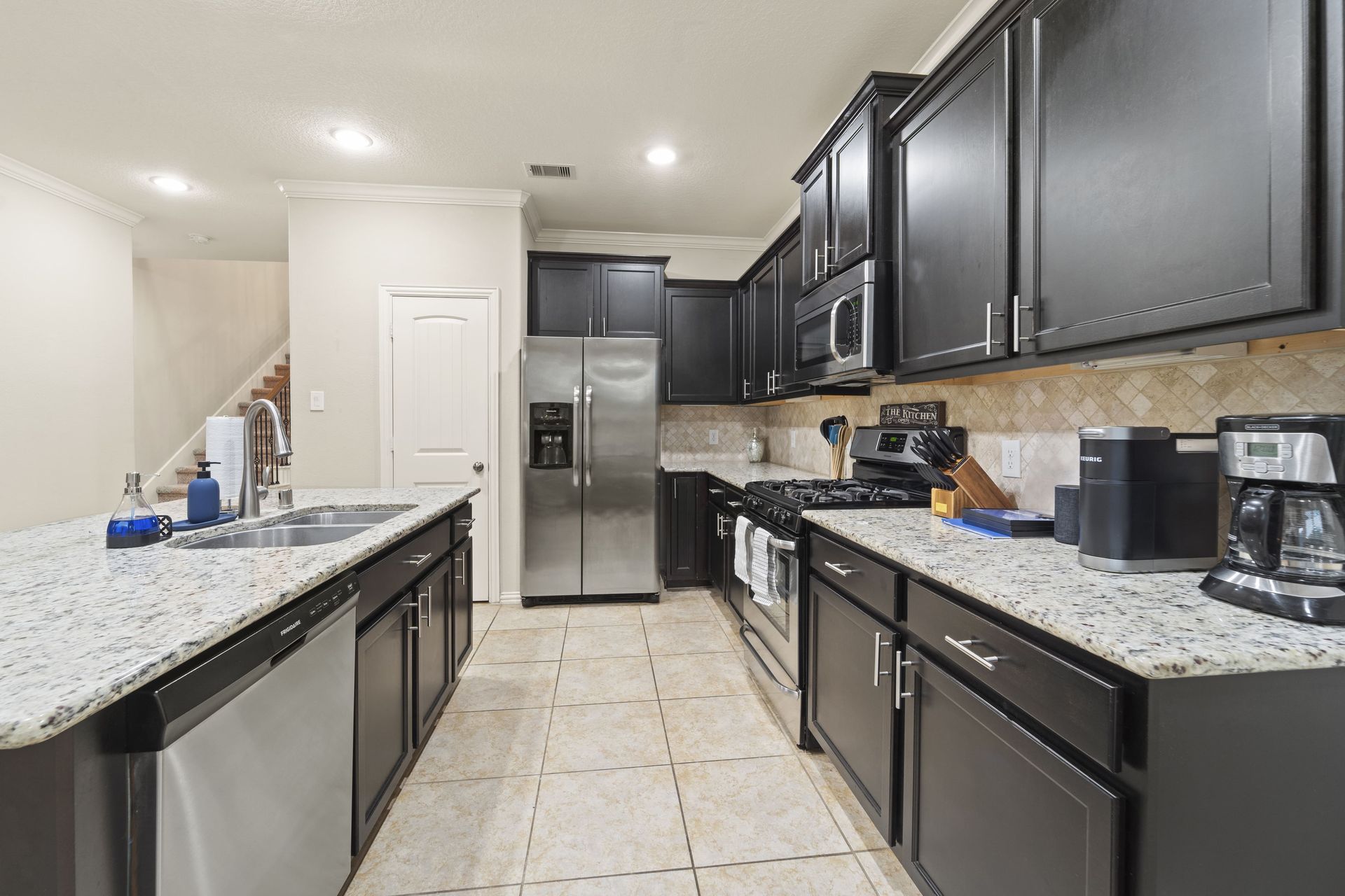 A kitchen with dark cabinets, granite countertops, stainless steel appliances, and a central island. Light-colored tile flooring is visible.