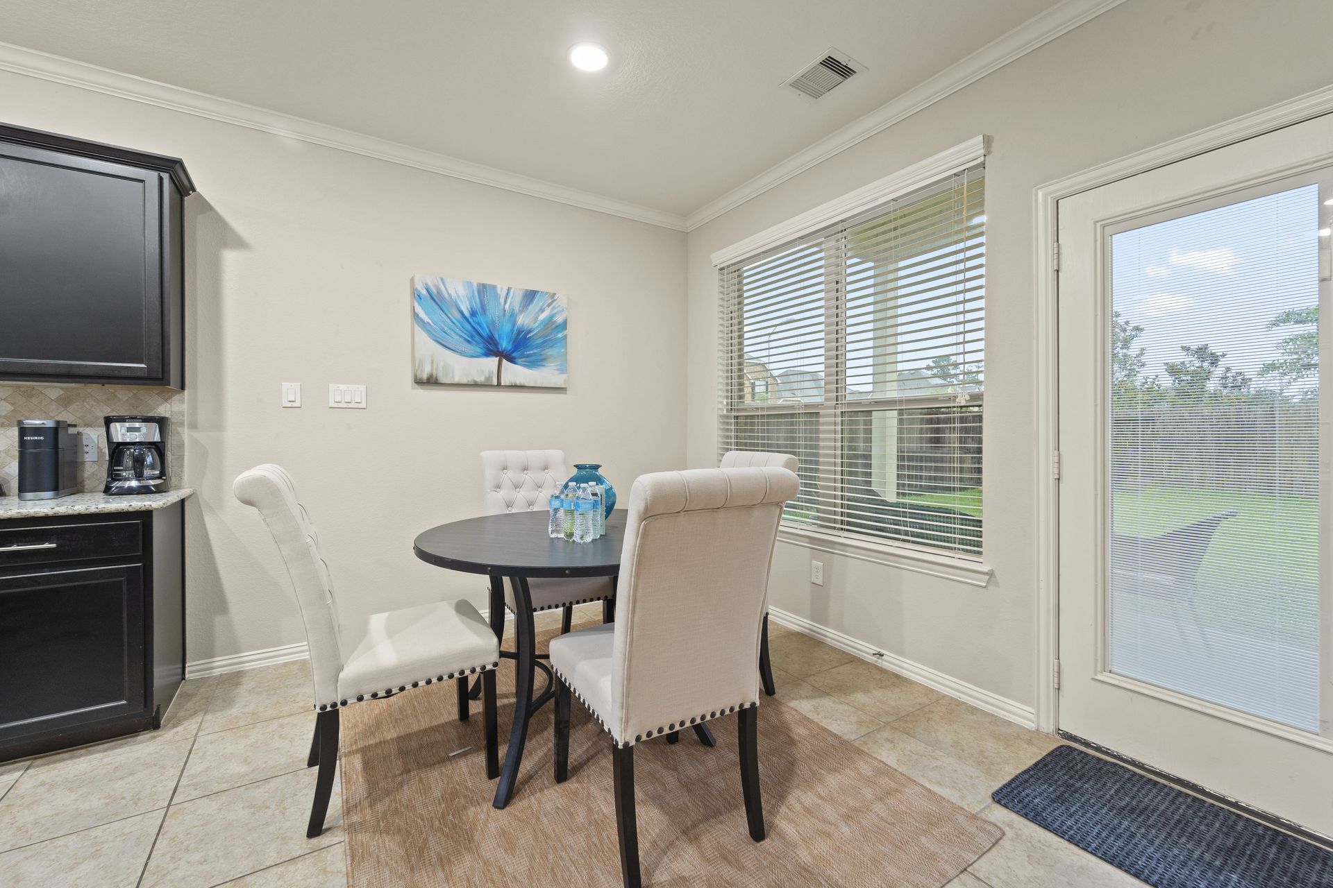 A dining area with a round table, four chairs, and a blue flower painting on the wall. A door and window with blinds provide outdoor views.