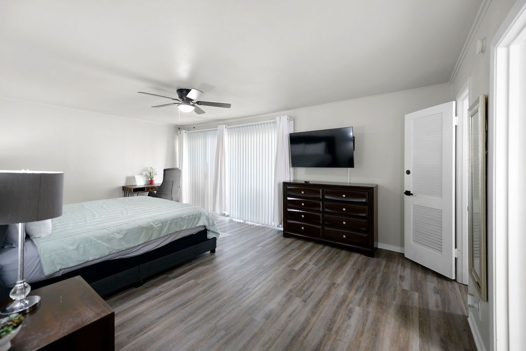 Spacious bedroom with a bed, dresser with a TV, curtains, and a door. Light wood-look floors and white walls.