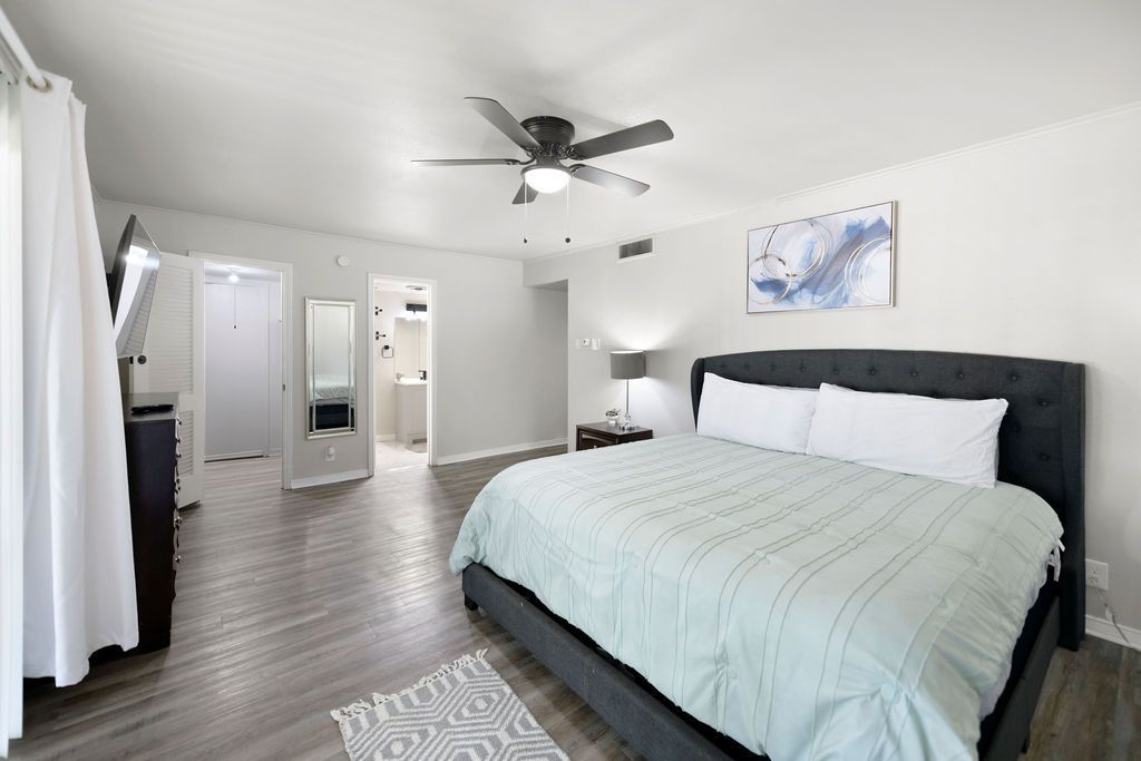 Spacious bedroom with a large bed, light-colored bedding, and a rug. There's a closet and bathroom visible in the background.